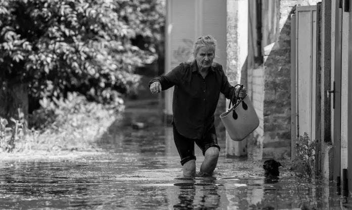 Black and white image of an elderly woman wading through a flooded street, holding a bucket and carefully navigating the water