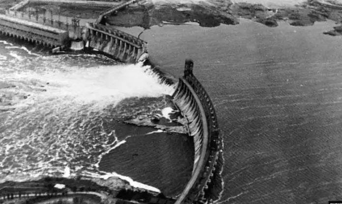 Black and white aerial photograph showing water forcefully spilling over a curved dam, illustrating the powerful movement and containment of water